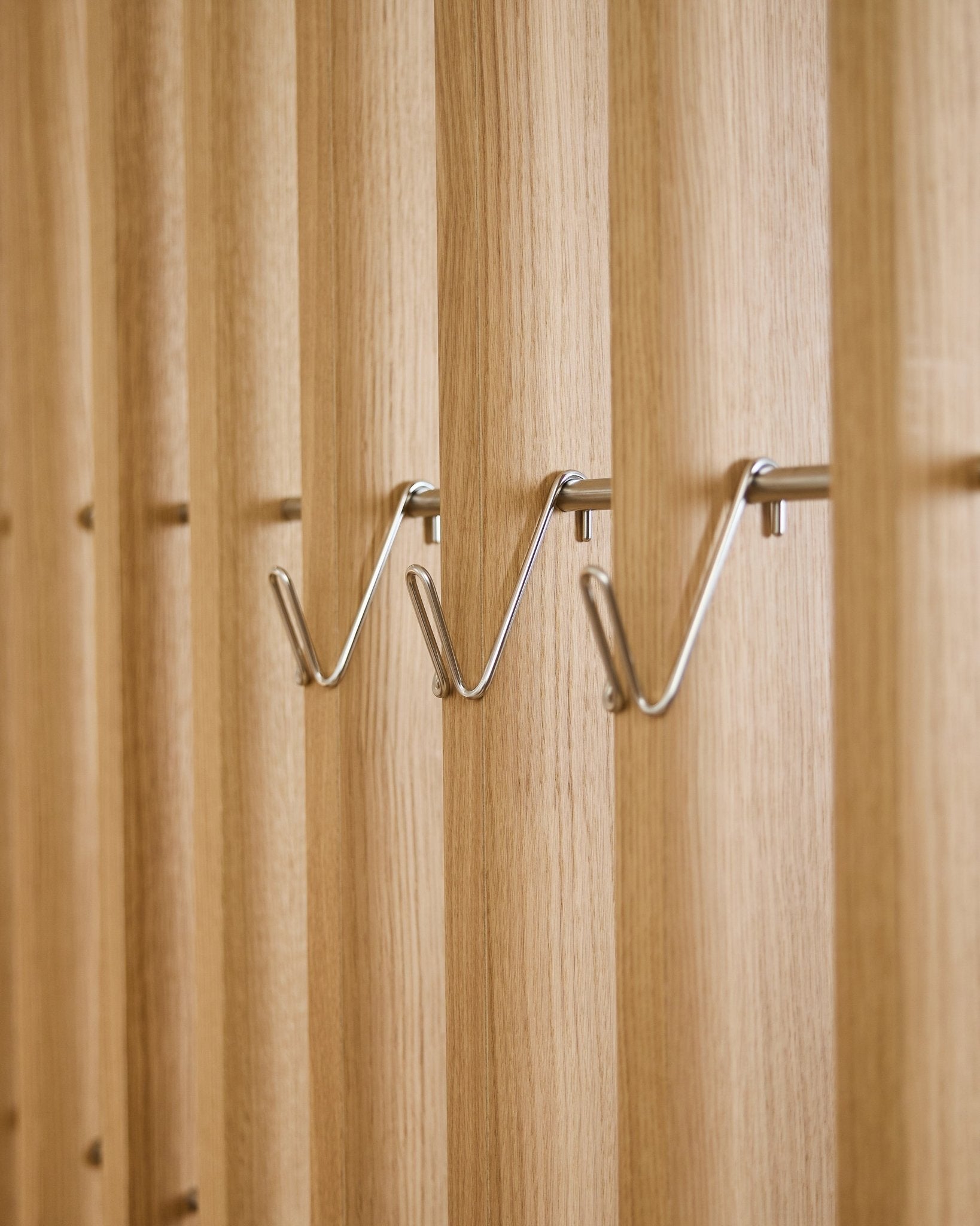 Tres accesorios Alva Wire Hook / BM1 de WoodUpp, cortos y de acero inoxidable steel, están montados de forma uniforme en una fila sobre un panel acústico de lamas de madera vertical.