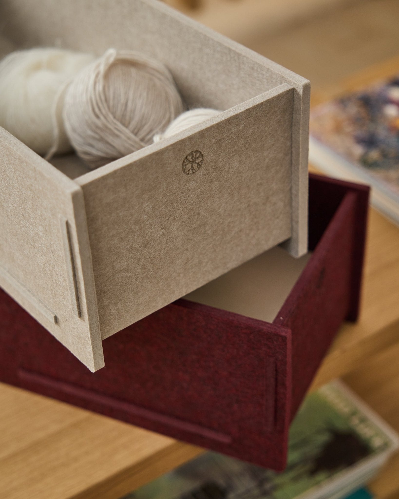 Una caja de almacenaje de fieltro PET beige llena de ovillos descansa sobre una WoodUpp Vigg Felt Box/BM2 cerrada en ox red, colocada sobre suelo de madera con libros al fondo.
