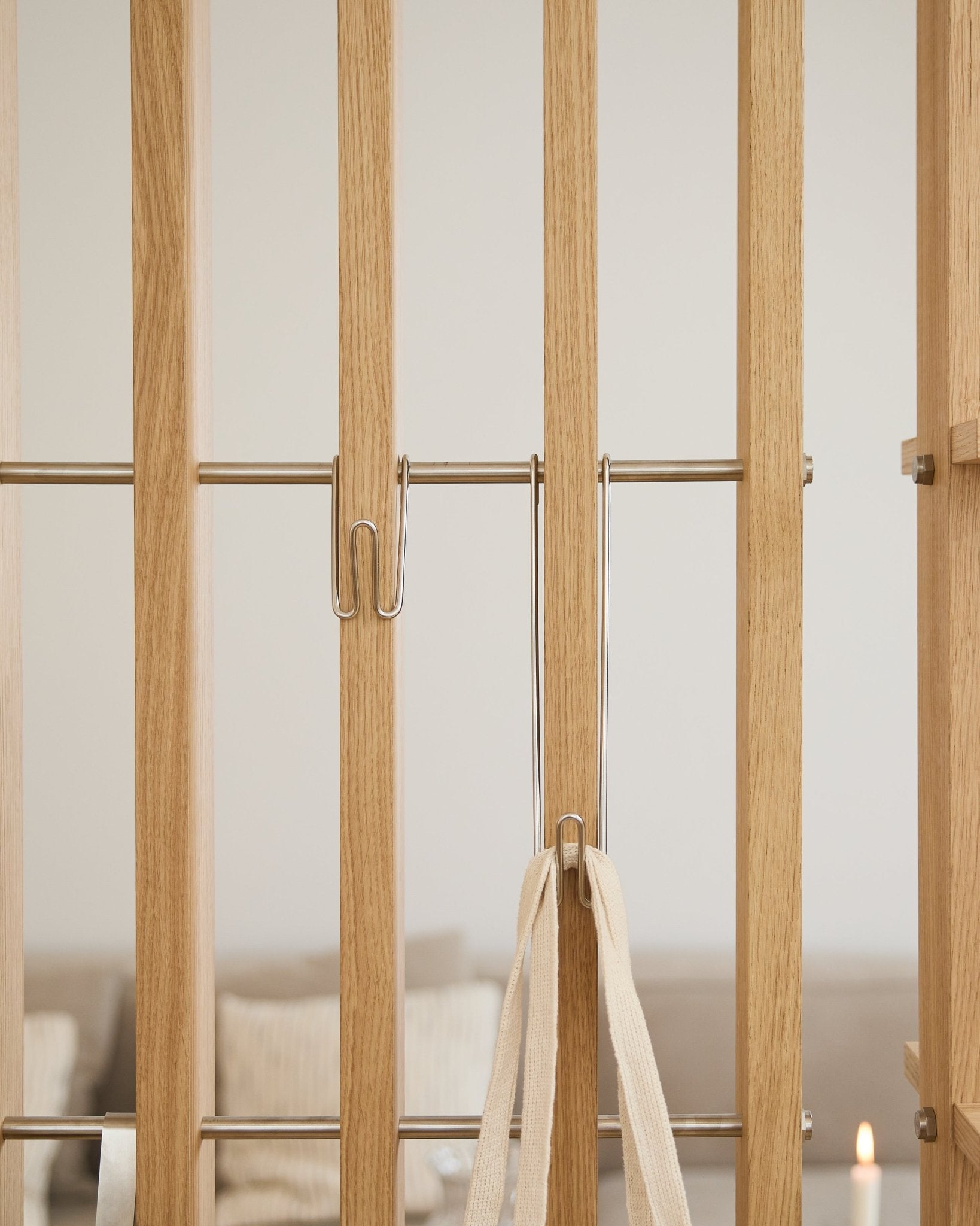 Primer plano de WoodUpp Alva | Wire Hook BM1 (acero inoxidable steel, largo, juego de 3) fijado a lamas claras Panel acústico con una correa blanca; sofá y vela desenfocados al fondo.