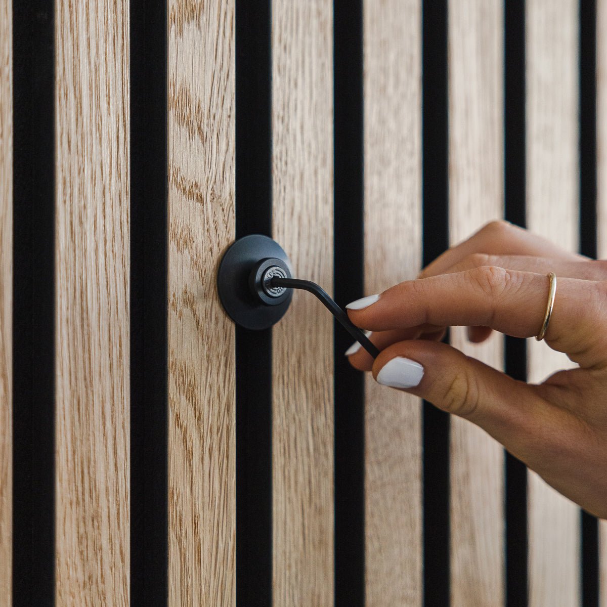 Una mano con esmalte de uñas blanco gira un gancho de aluminio Dupps | Heavy black de WoodUpp sobre un panel acústico de madera vertical Akupanel.