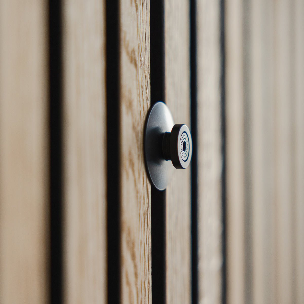 Primer plano de la mirilla Dupps | Heavy de WoodUpp en aluminio black, instalada en una puerta de madera ranurada con lamas acústicas Akupanel, con la lente enfocada.