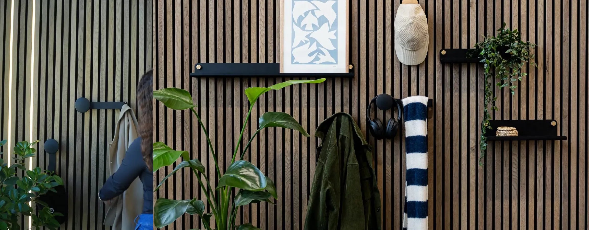 Un hombre con chaqueta azul pasa junto a un muro de lamas de madera acústica Akupanel nórdica con plantas integradas, estanterías, perchas, auriculares y una toalla de rayas enrollada.