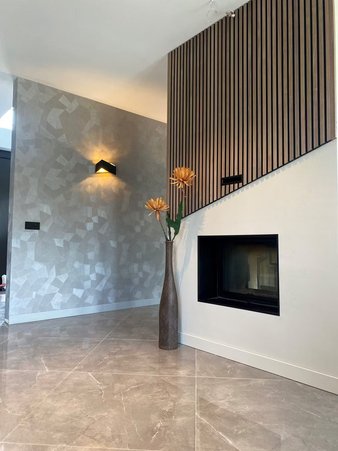 Moderno salón con papel pintado de motivos geométricos, paneles acústicos de madera de Akupanel, chimenea de pared y un jarrón alto de flores naranjas.