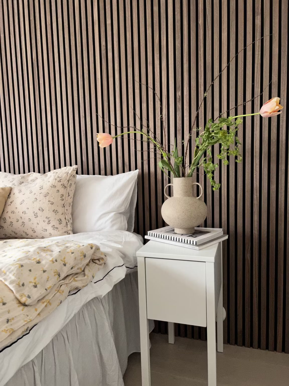 Un dormitorio con una cama blanca con ropa de cama floral y un jarrón de flores, con una pared nórdica de listones acústicos de madera Akupanel detrás de la cama.