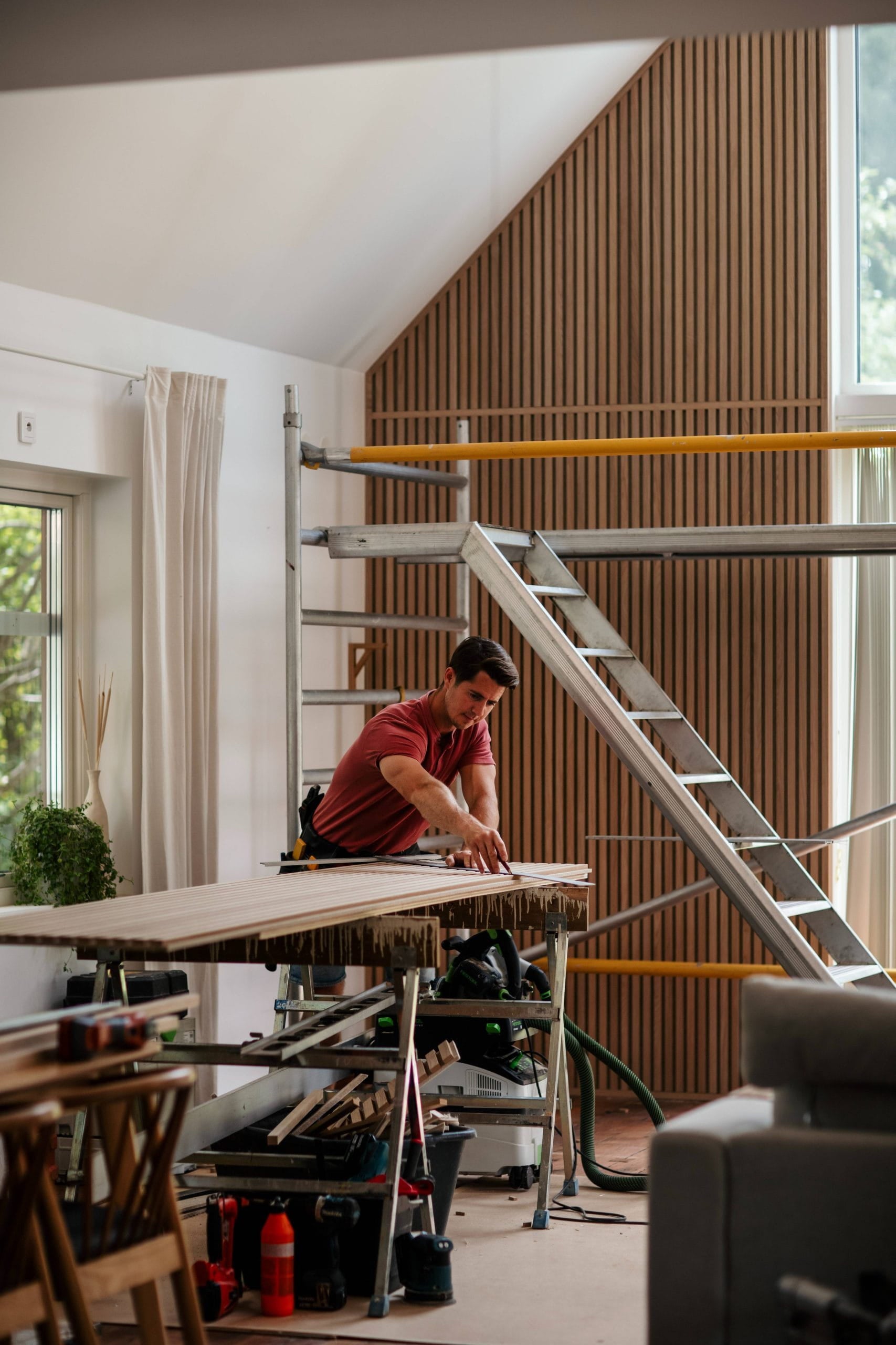 Un hombre mide un tablón de madera sobre un banco de trabajo en una sala moderna, parcialmente renovada, con andamios y grandes ventanales, lo que sugiere la instalación de paneles acústicos.