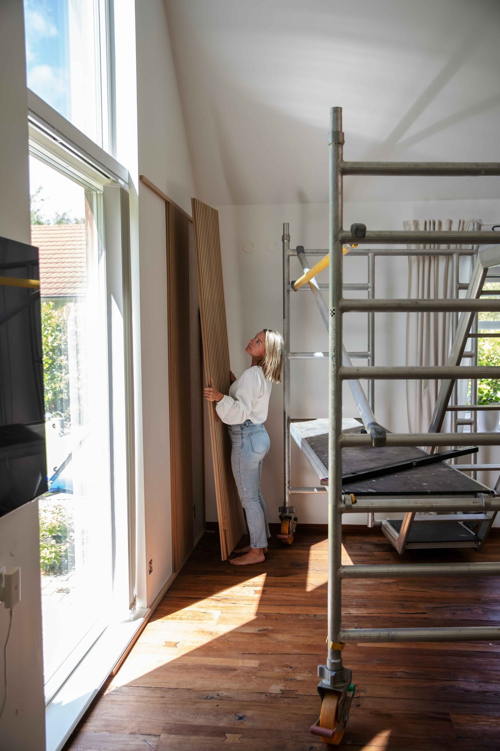 Una mujer se encuentra en una habitación iluminada por el sol, con grandes ventanales y andamios, sosteniendo un panel de madera acústica Akupanel sobre un suelo de madera pulida.