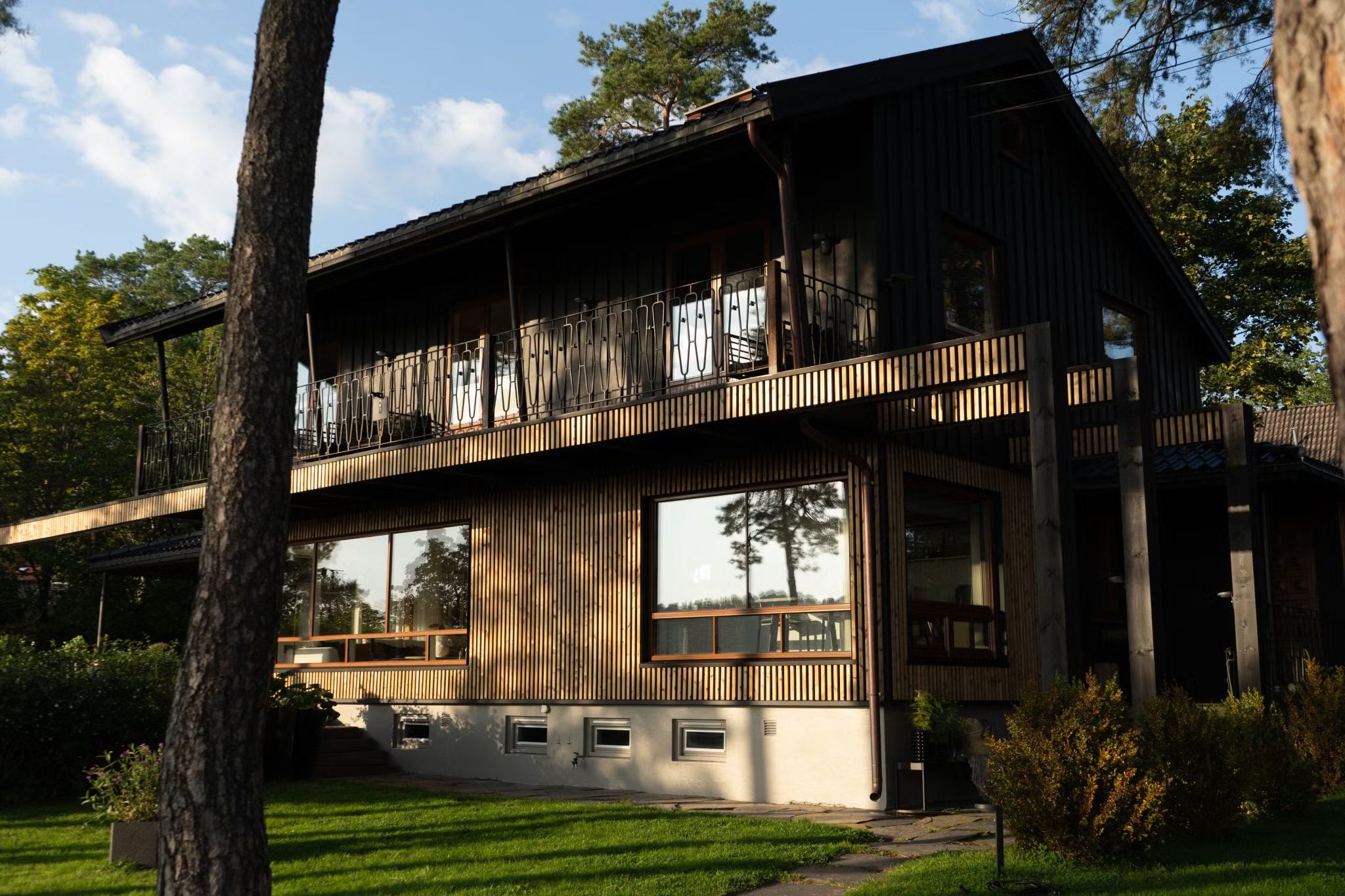 Moderna casa de dos plantas con grandes ventanales y paneles de madera oscura Aluwood en la fachada, rodeada de árboles y un verde césped bajo la luz del sol.