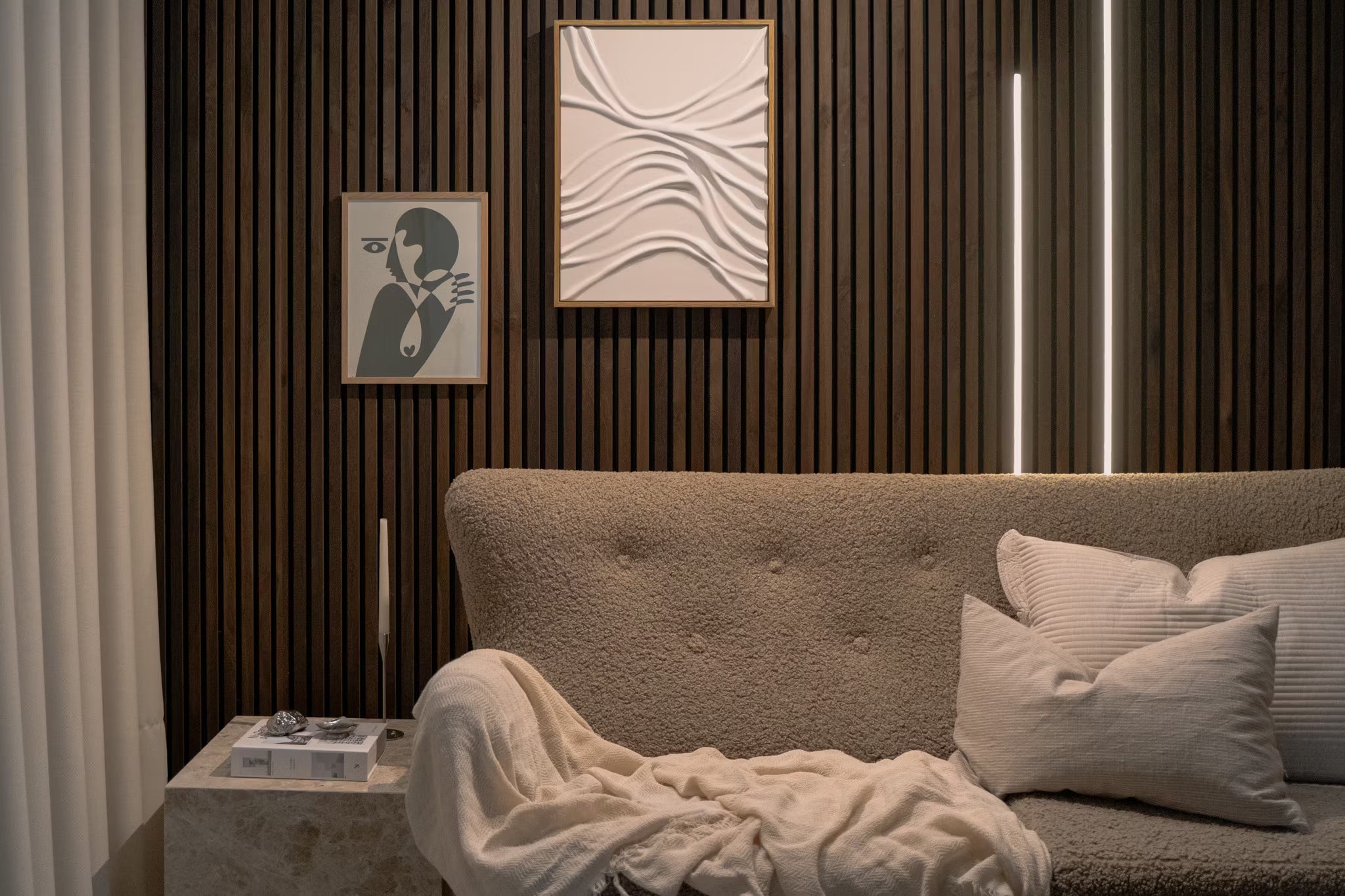 Moderno salón con sofá beige texturizado, cojines blancos, arte abstracto en paneles acústicos y pared vertical de listones de madera Akupanel con suave iluminación ambiental.