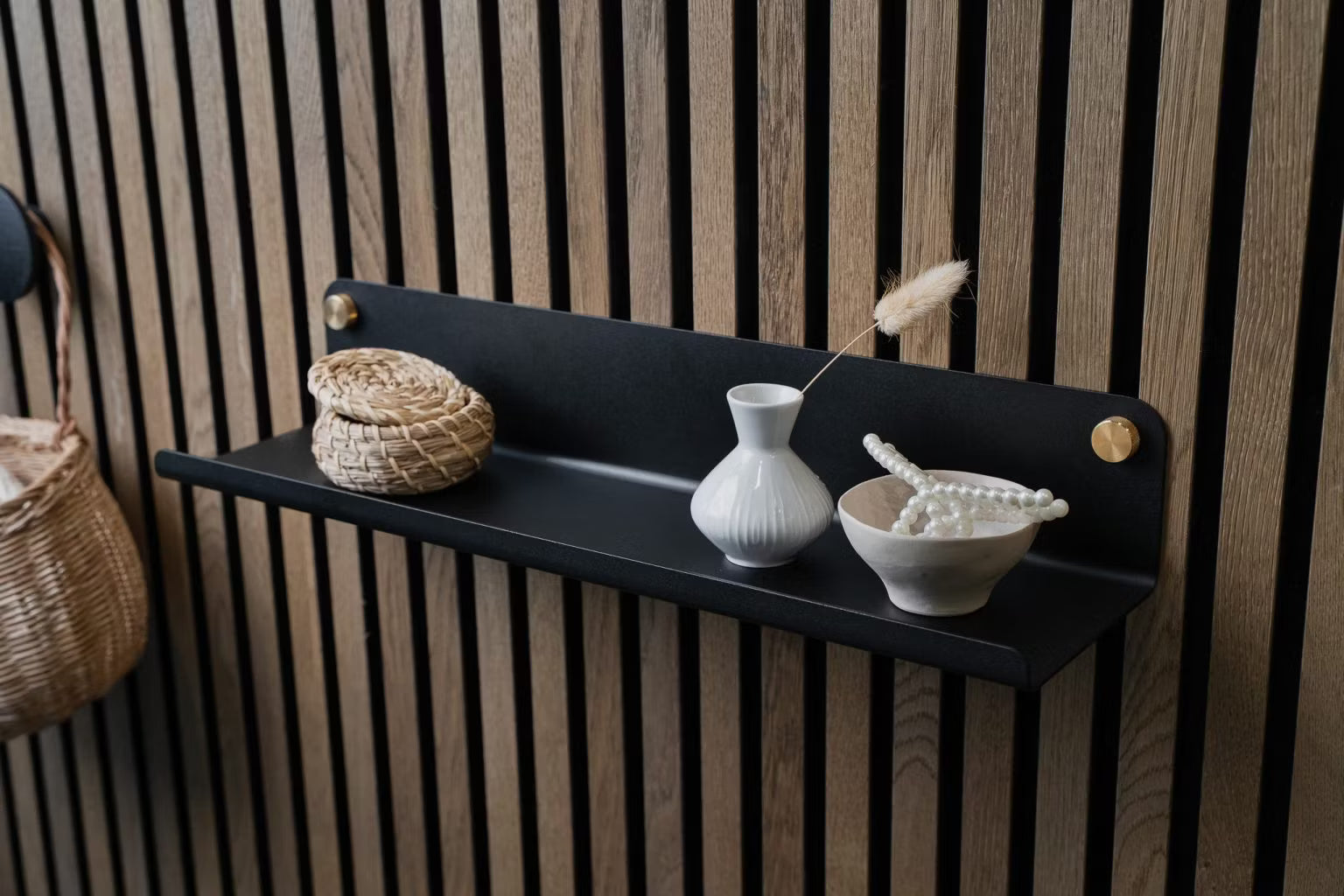Una estantería de pared negra con una pequeña cesta de mimbre, un jarrón blanco y un cuenco de perlas de cerámica montados sobre un panel nórdico de listones acústicos de madera.