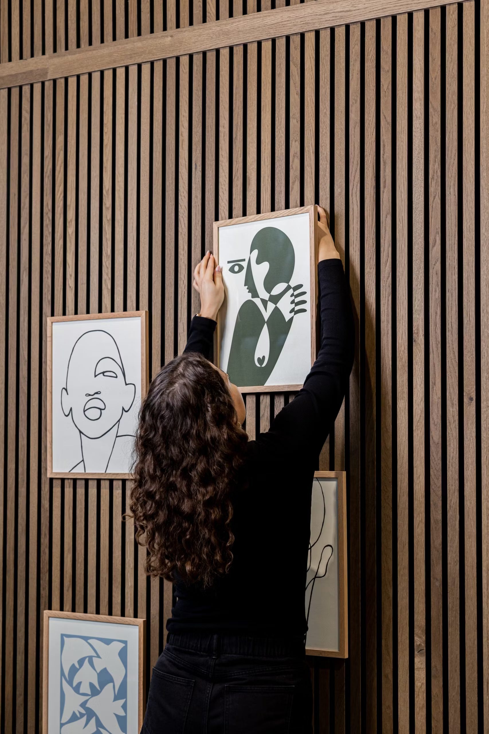 Una persona con el pelo rizado cuelga un accesorio WoodUpp® CREATE DUPPs en un panel acústico de listones de madera, rodeado de arte minimalista y decoración moderna.