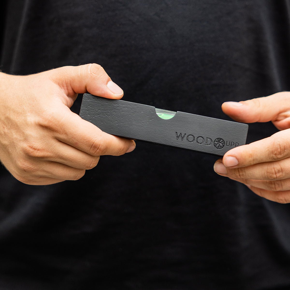 Una persona con una camiseta black sostiene un nivel de burbuja WoodUpp black (15 x 3,2 cm), diseñado para una instalación precisa de paneles acústicos.