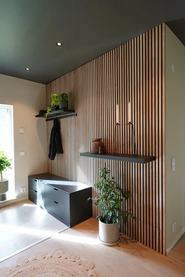 Entrada moderna con panel WoodUpp de listones verticales en madera natural, estantes flotantes negros WoodUpp de acabado mate, zapatero/banqueta baja con cajones negros, perchero superior con repisa, plantas en macetas y candelero. Luz natural realza textura y líneas verticales del revestimiento WoodUpp.