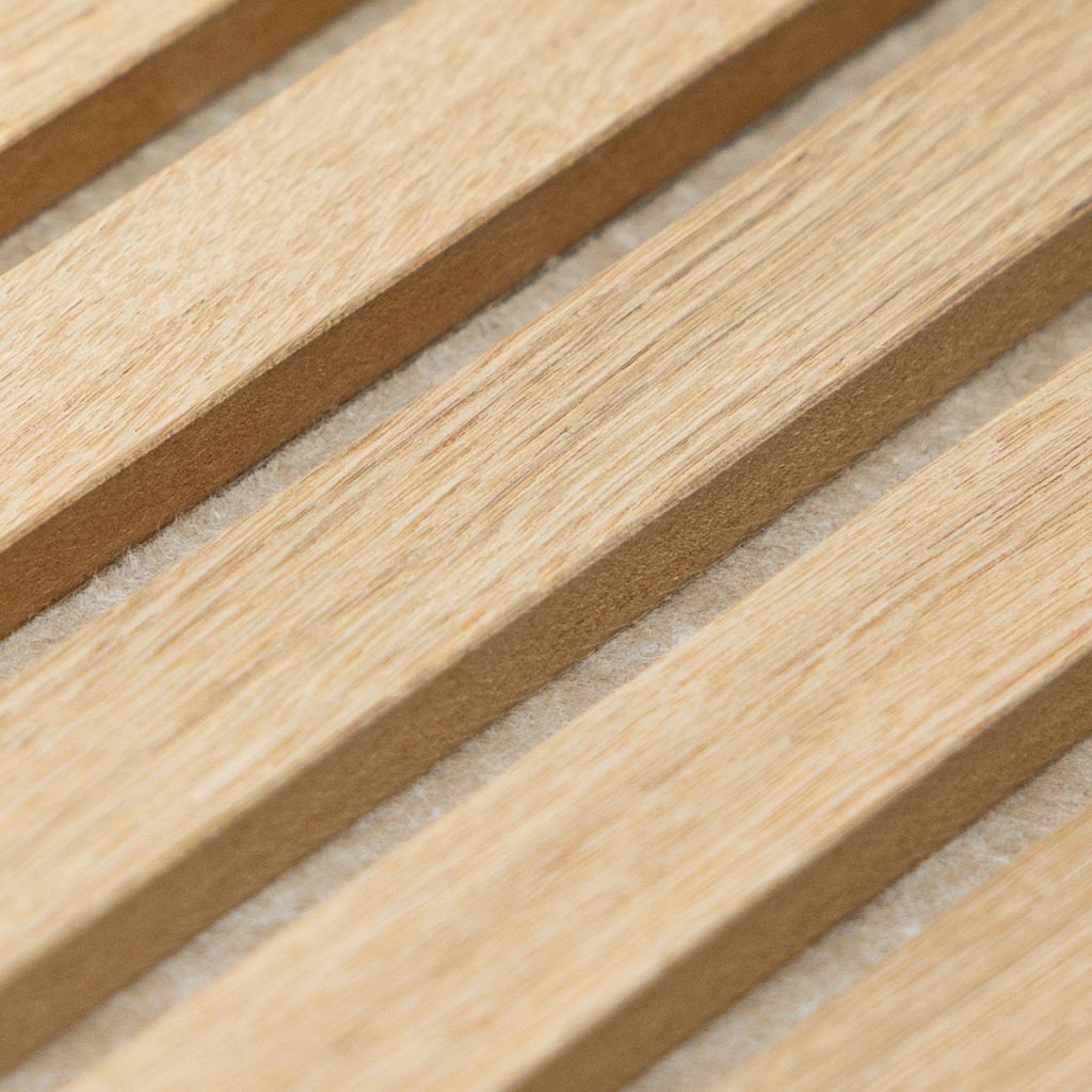 Primer plano de un panel acústico WoodUpp formado por lamas verticales de madera clara con vetas visibles, montadas sobre un respaldo textil de fieltro absorbente gris claro; las lamas tienen acabado natural y separaciones regulares que dejan ver el material aislante entre ellas.