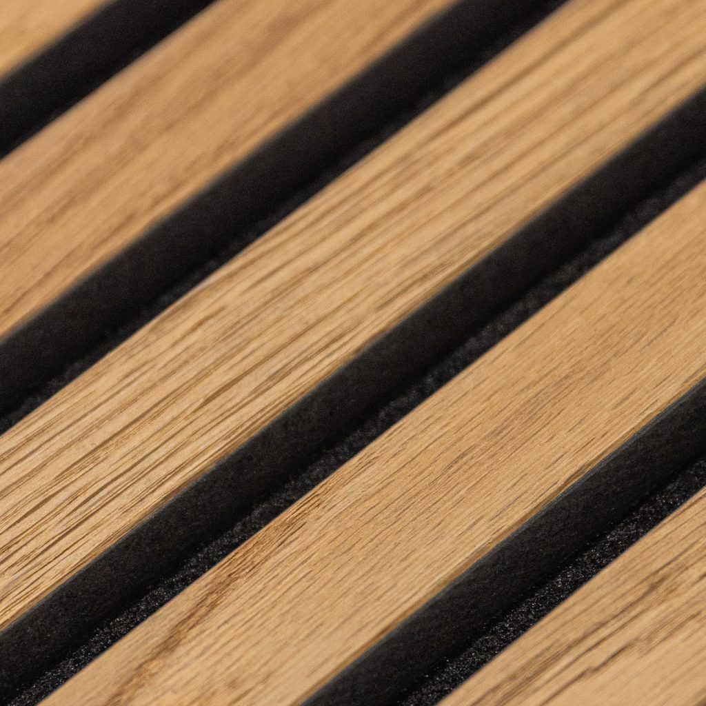 Primer plano de un panel acústico WoodUpp: finas lamas de madera clara con vetas visibles, acabado mate, dispuestas en diagonal y separadas por tiras negras de espuma o fieltro que crean ranuras profundas. Detalle muestra textura natural y contraste entre madera cálida y juntas oscuras.
