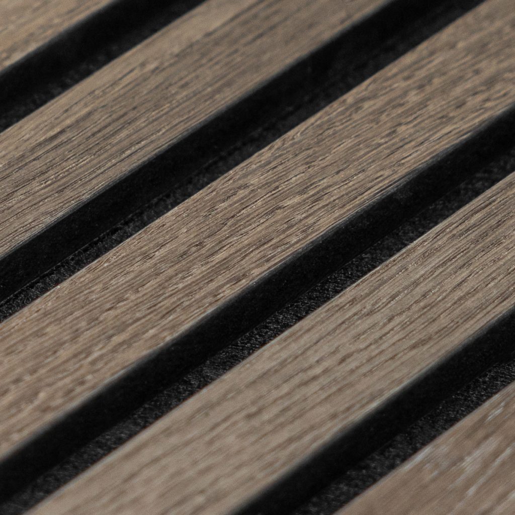 Primer plano de panel acústico WoodUpp: listones de madera clara con vetas visibles y acabado cepillado, dispuestos en franjas diagonales. Entre los listones se aprecian ranuras rellenas de material negro absorbente (fieltro/espuma), creando contraste y textura en la superficie del producto.