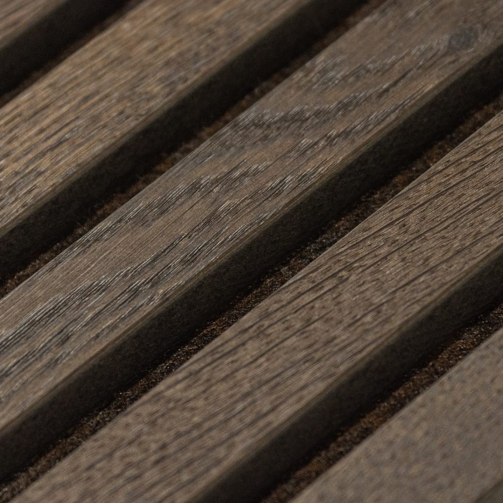 Detalle macro de panel acústico WoodUpp: listones de madera oscura colocados en diagonal con veta marcada, separados por ranuras rellenas de fieltro/espuma marrón. Producto WoodUpp de lamas acústicas que combinan textura natural de la madera con aislamiento sonoro y acabado mate elegante.
