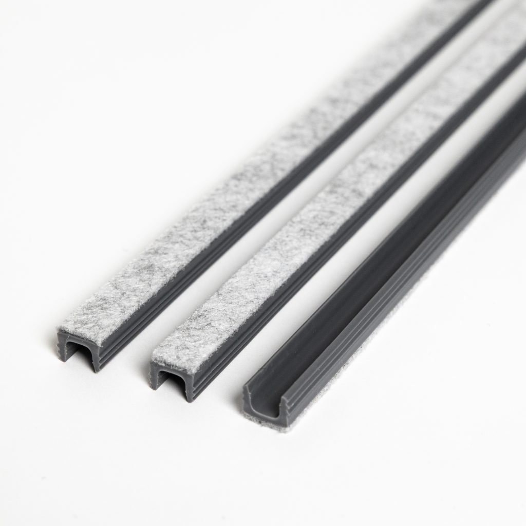 Imagen de WoodUpp que muestra tres perfiles en U de plástico gris con tiras de fieltro gris claro adheridas; dos perfiles más estrechos y uno más ancho, colocados en diagonal sobre fondo blanco. Productos aparentes: burletes o guías de fieltro para puertas y muebles, de plástico y fieltro.