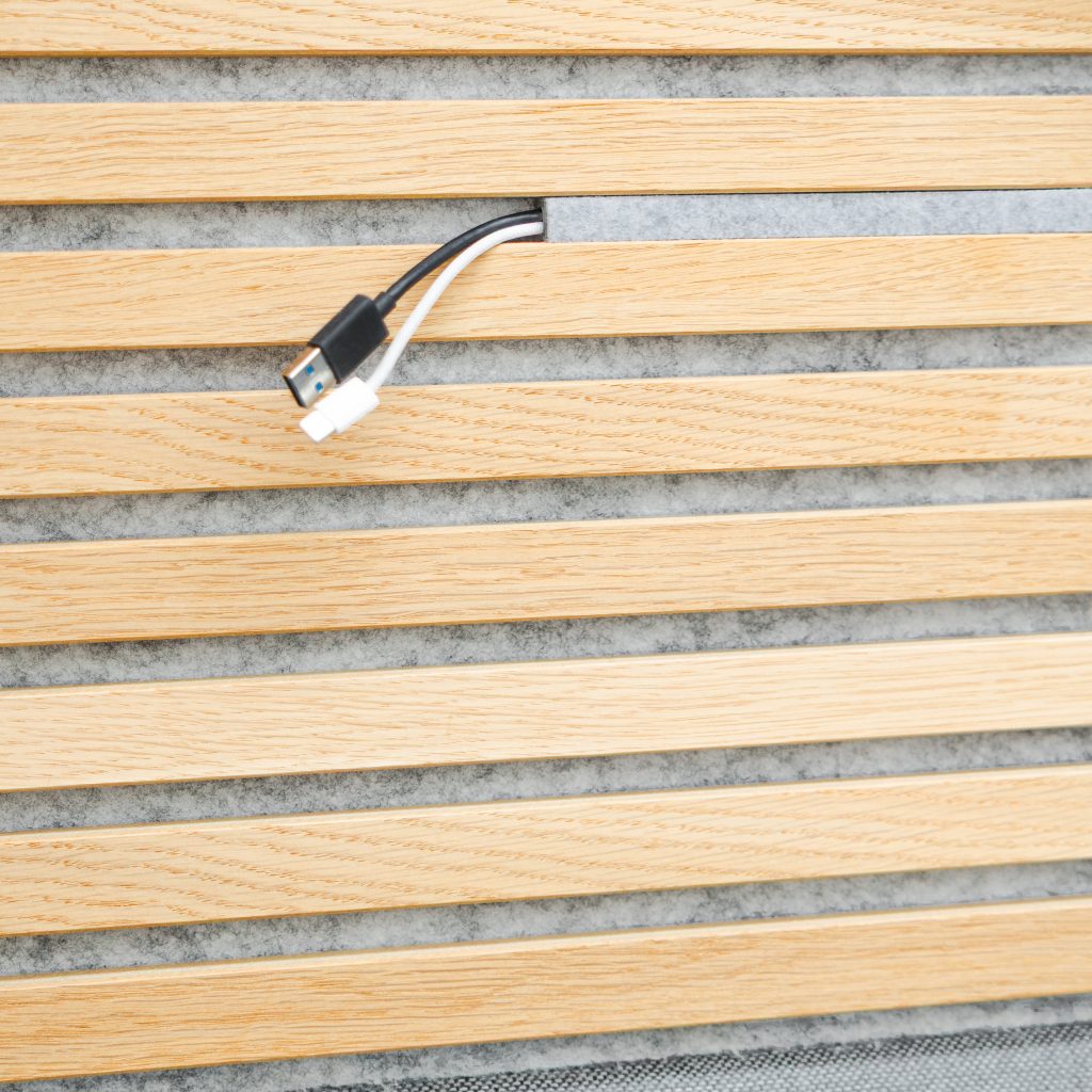 Panel WoodUpp de lamas de madera clara (roble) con respaldo de fieltro acústico gris; ranura horizontal para gestión de cables; de la abertura salen dos cables: uno USB‑A negro y otro blanco de carga, mostrando el acabado y la funcionalidad del producto WoodUpp.