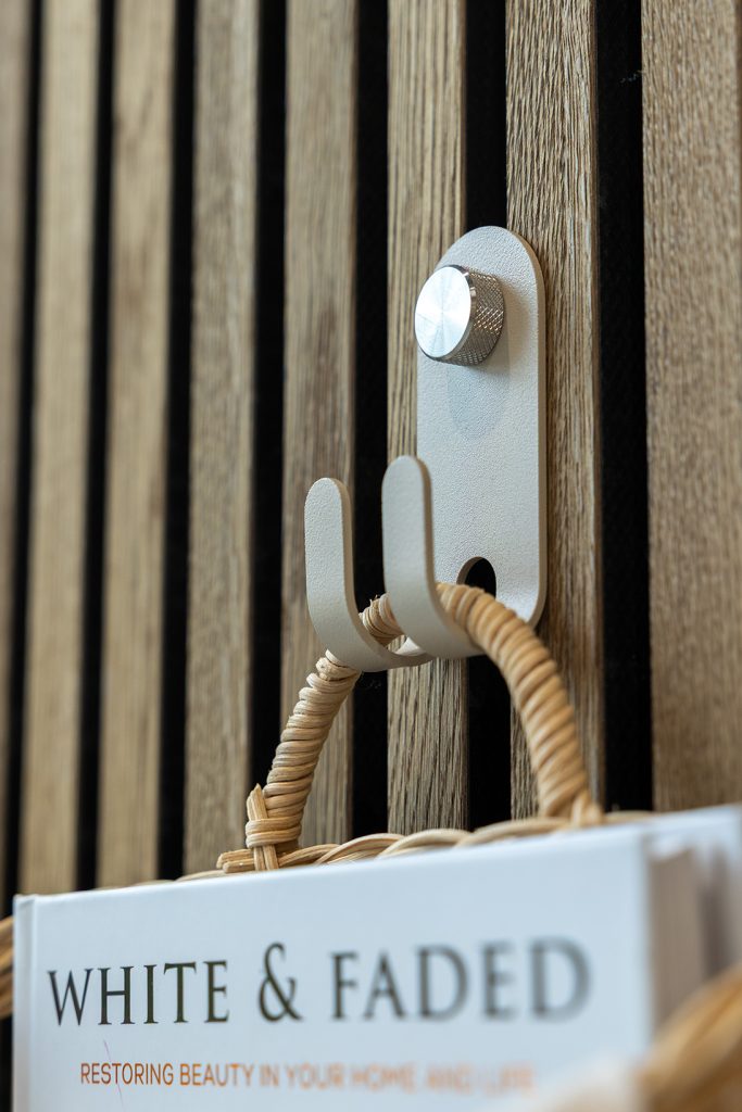 Lamas verticales de madera WoodUpp en acabado roble sobre fondo oscuro, acompañadas por un gancho doble metálico WoodUpp color crema fijado con un perno estriado; del gancho cuelga el asa trenzada de una cesta y se aprecia la esquina del libro "White & Faded".