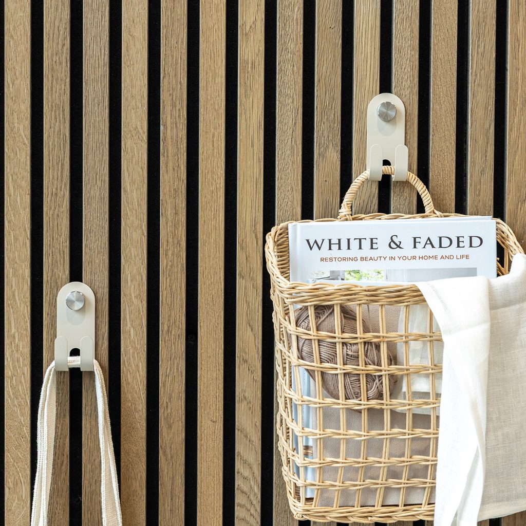 Panel de lamas de madera con dos colgadores metálicos crema de WoodUpp, de gancho doble y remache circular; uno sostiene un bolso de tela y el otro sujeta una cesta de mimbre colgada con la revista "White & Faded", un ovillo de lana y una tela clara.