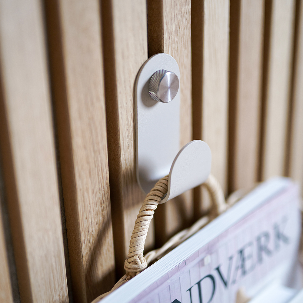 Colgador modular WoodUpp: gancho metálico blanco-crema fijado al panel de lamas de madera mediante un botón cilíndrico de metal estriado, sosteniendo una cesta de mimbre con revistas. Imagen en primer plano muestra detalle del diseño minimalista y la fijación robusta del producto WoodUpp.