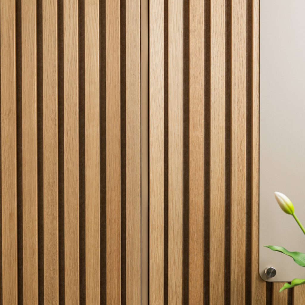 Panel mural vertical de lamas de madera de WoodUpp, acabado roble claro con ranuras oscuras entre listones, mostrando un panel acústico decorativo. A la derecha aparece un tablero claro sujeto con un tornillo metálico y una yema de flor verde, destacando textura y diseño.