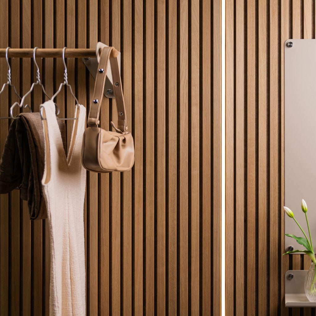 Panel WoodUpp de listones verticales de roble claro formando una pared texturizada; perchero WoodUpp con barra de madera y soportes metálicos que sostiene abrigos, perchas y un bolso beige; estante pequeño y espejo fijados a la pared con un jarrón y flores.