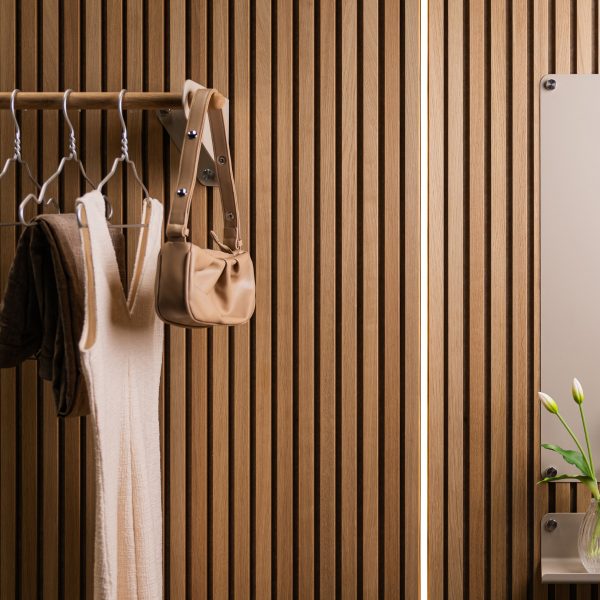 Panel WoodUpp de listones verticales de roble claro formando una pared texturizada; perchero WoodUpp con barra de madera y soportes metálicos que sostiene abrigos, perchas y un bolso beige; estante pequeño y espejo fijados a la pared con un jarrón y flores.