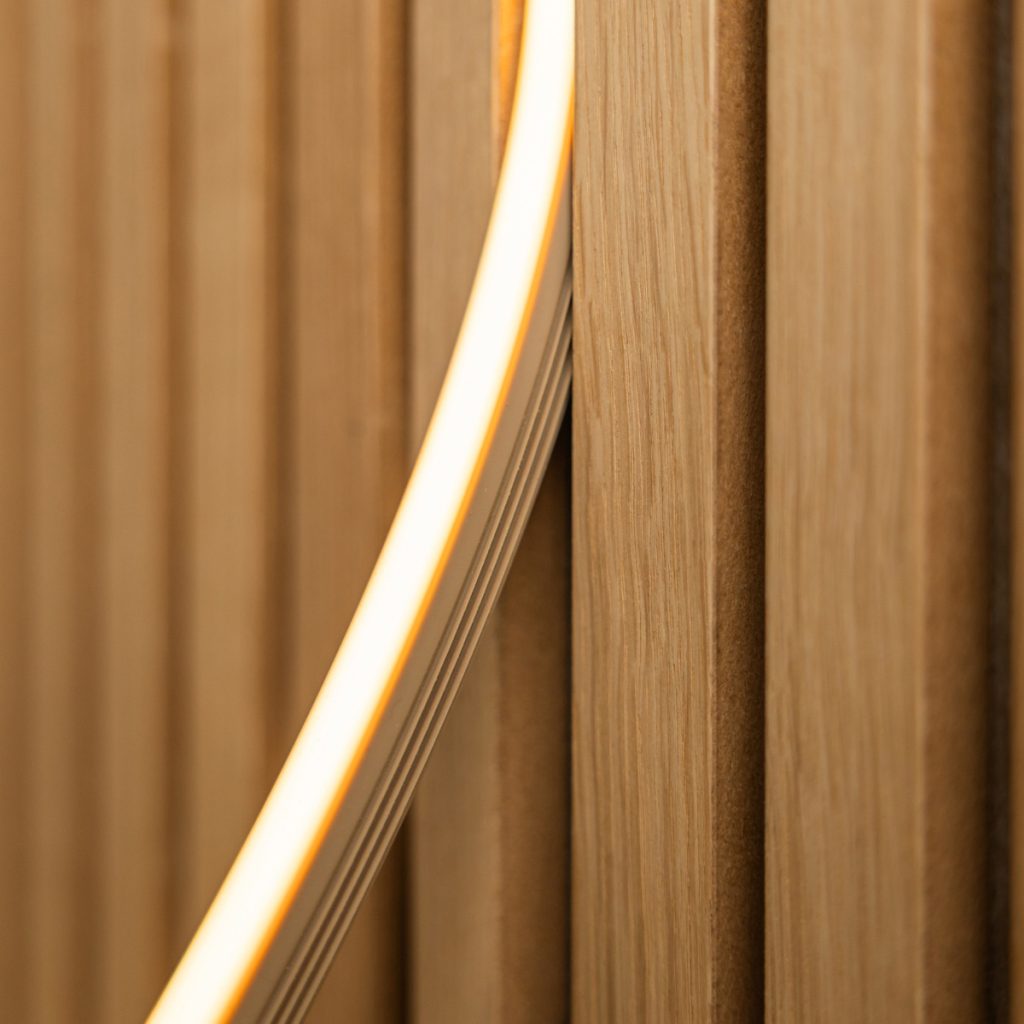 Detalle de paneles verticales ranurados de madera clara WoodUpp, con veta visible y listones perfilados, atravesados por una tira LED flexible iluminada en tono cálido, mostrando la textura y el acabado natural de los revestimientos WoodUpp y su luminaria integrada.