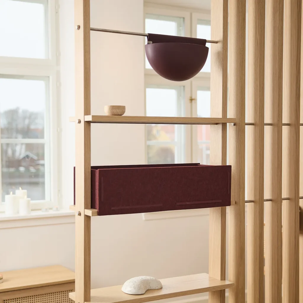 Estantería modular WoodUpp de listones y baldas de roble claro, usada como biombo; con un cajón organizador de fieltro burdeos, una maceta colgante burdeos, un pequeño cuenco de madera sobre la balda y una piedra decorativa blanca sobre la estantería.
