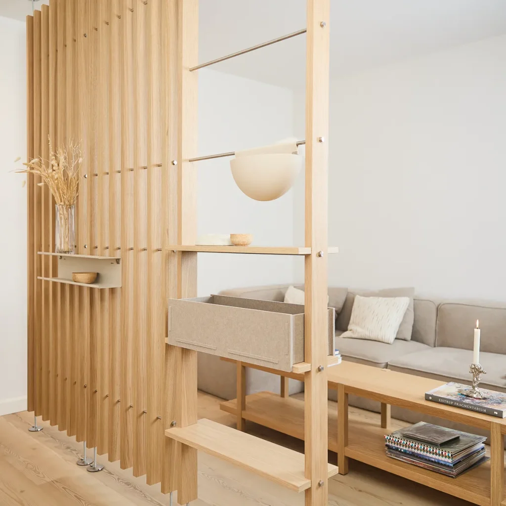 Separador WoodUpp de lamas verticales en madera clara con estantes modulares, cajón de fieltro deslizante y bandejas auxiliares fijadas a varillas metálicas. Incluye repisas abiertas, una lámpara colgante y un estante pequeño con cuenco; organiza y divide el salón con estilo minimalista.