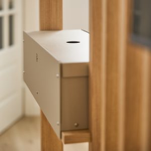 Detalle de un producto WoodUpp: caja metálica beige montada en una estructura de listones de madera natural. Caja rectangular con cierre superior y orificio circular, tornillos visibles y pequeño adorno perforado lateral; diseño minimalista para organización y gestión de cables o accesorios.