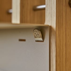 Primer plano de una instalación WoodUpp: un gancho metálico con logotipo circular sobre un panel metálico, montado en un marco de madera clara tipo roble. Se aprecian ranuras y una barra metálica desenfocada en el fondo, mostrando detalle y acabado de los productos WoodUpp.