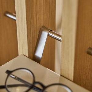 Detalle de madera clara WoodUpp con listones verticales y un colgador metálico abatible de acero cepillado; se aprecia una barra tubular horizontal y una repisa blanca inferior con gafas. Fotografía muestra acabado natural del roble y el diseño minimalista de los productos WoodUpp.