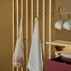 Panel listonado modular de madera roble de WoodUpp con rieles y ganchos metálicos; toalla de rayas y bolsa de red colgadas, estante con cuencos y riel para tazas con tazas blancas; cajón organizador de fieltro burdeos insertado en la estructura.