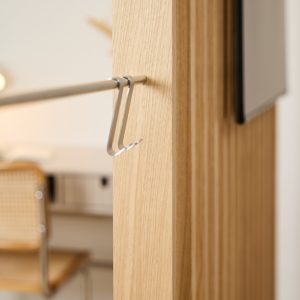 Detalle WoodUpp: panel vertical de madera clara tipo roble con listones, una barra metálica delgada instalada en el panel y dos ganchos en S de acero inoxidable colgados en la barra, diseñados para colgar accesorios con acabado mate y aspecto minimalista.