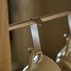 Primer plano de un colgador WoodUpp: una abrazadera metálica de acabado cepillado con logo grabado sujeta una barra cromada y sostiene una taza de cerámica colgada en un gancho metálico. Detalle del clip WoodUpp, diseño minimalista y acabado inoxidable y elegante.