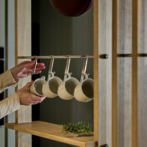 Estantería modular de WoodUpp en madera clara con barra metálica para colgar, donde se ven ganchos metálicos WoodUpp sosteniendo cuatro tazas de cerámica blanca. Unas manos colocan una taza; en la balda hay hierbas frescas. Diseño minimalista y funcional para cocina.