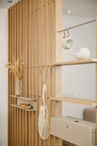 Panel divisor de lamas de madera de roble de WoodUpp con estantes flotantes y cajón metálico modular beige, barra colgante y ganchos metálicos WoodUpp; muestra taza colgada, jarrón con flores secas, cuencos de madera, vela y bolsa de red colgante, diseño minimalista y funcional.