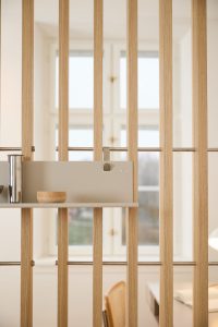 Separador WoodUpp formado por listones verticales de madera clara (roble) y barras metálicas, con un panel estante modular gris de WoodUpp sujeto con clips. En el estante hay un cuenco de madera y una jarra metálica, destacando el diseño minimalista y funcional de WoodUpp.