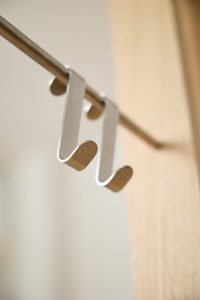 Dos ganchos colgadores metálicos plateados de WoodUpp, de diseño en U plana, colgados sobre una barra metálica; muestran un logotipo circular grabado en los extremos. Foto en primer plano con fondo difuminado y borde de puerta de madera clara visible.