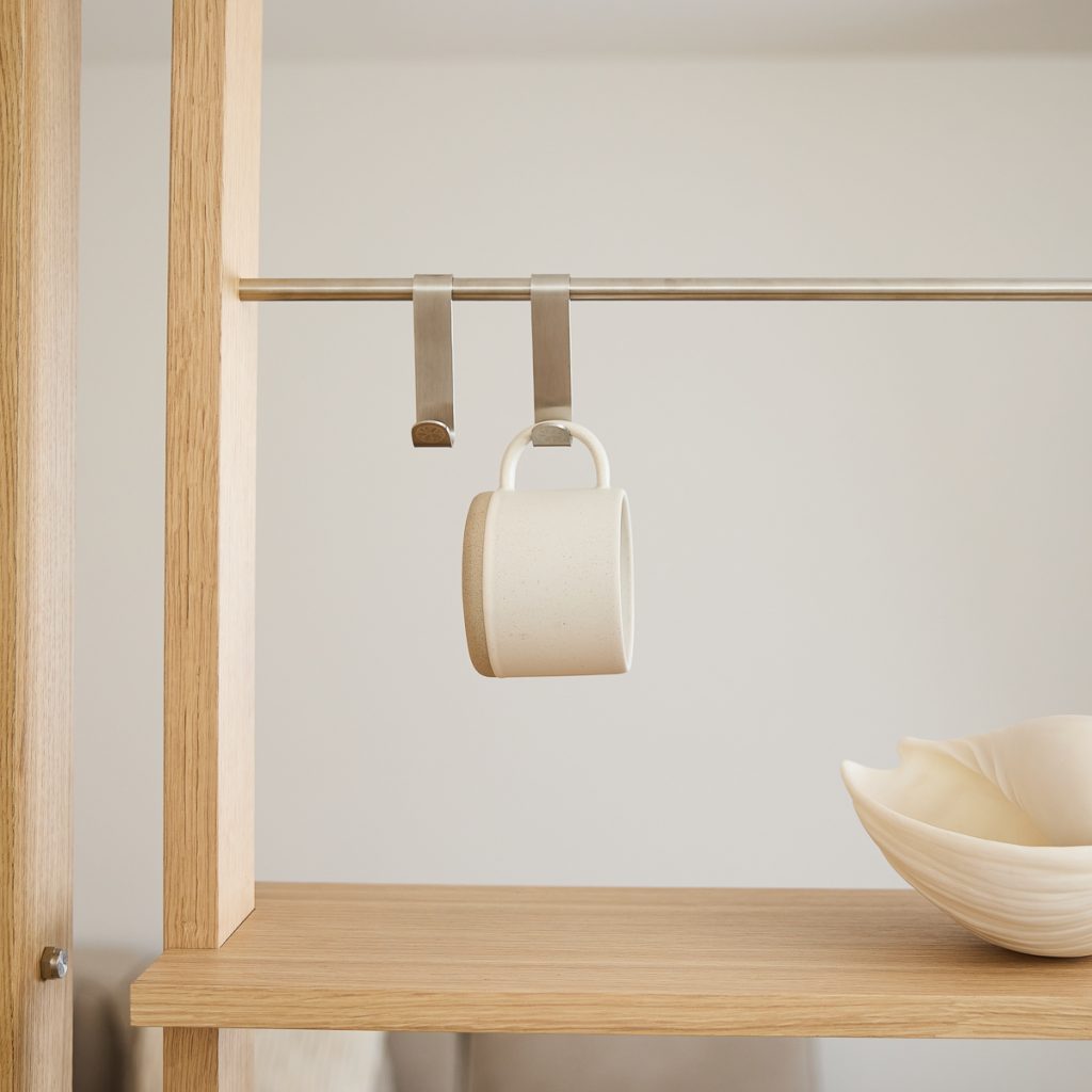 Estantería de madera natural WoodUpp con barra metálica y dos ganchos colgantes de acero inoxidable WoodUpp; uno sostiene una taza cerámica beige suspendida y sobre la balda hay un cuenco de cerámica claro, composición minimalista, elegante y con detalles pulidos.